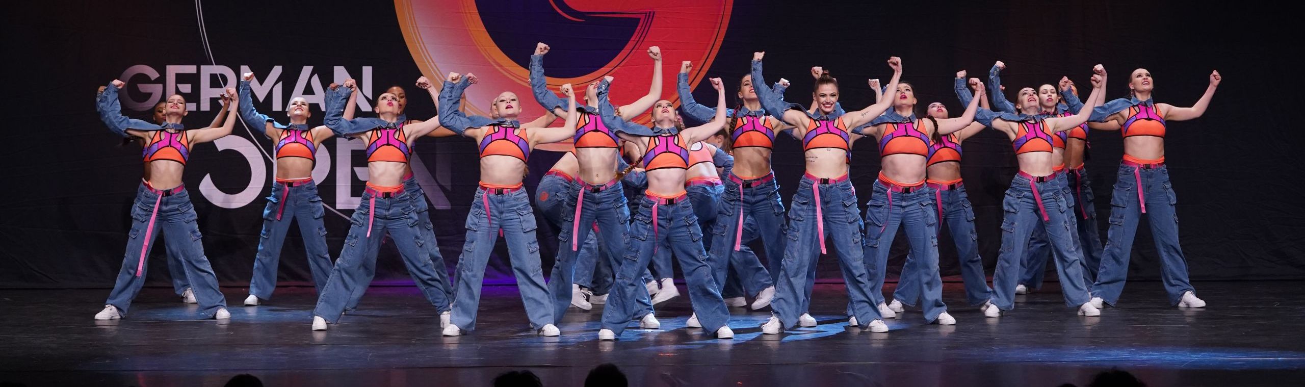 Tanzformation mit Jeans und leuchtenden Oberteilen auf der Bühne